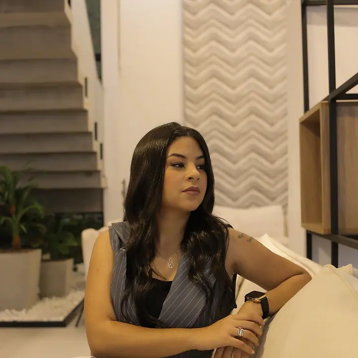 Dra. Akemy Rande de Sousa sentada em um sofá branco, de colete risca de giz, olhando para a câmera