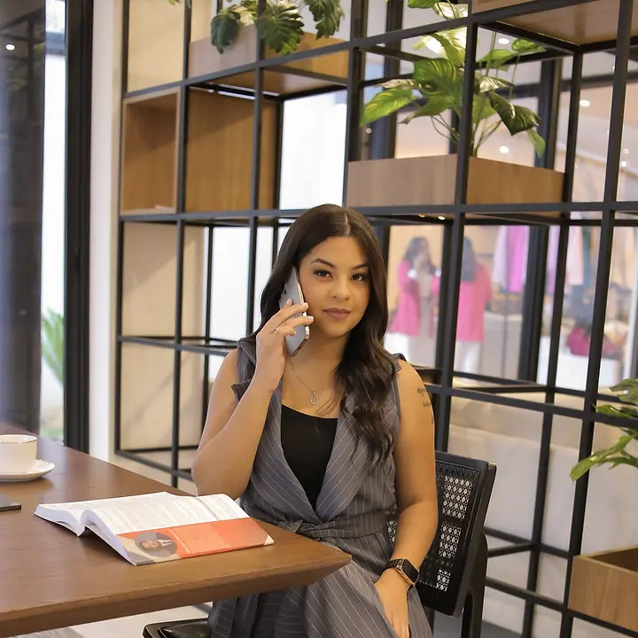 Dra. Akemy Rande de Sousa ao telefone, sentada à mesa de trabalho, de colete e calça risca de giz